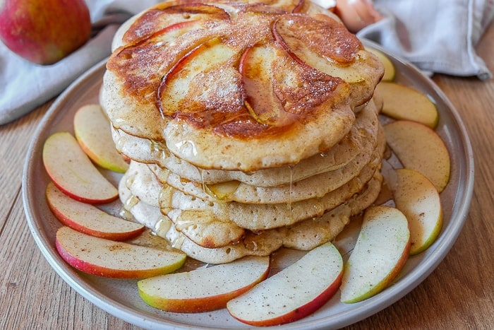 stack of apple pancakes on plate with sliced apples around on wooden table