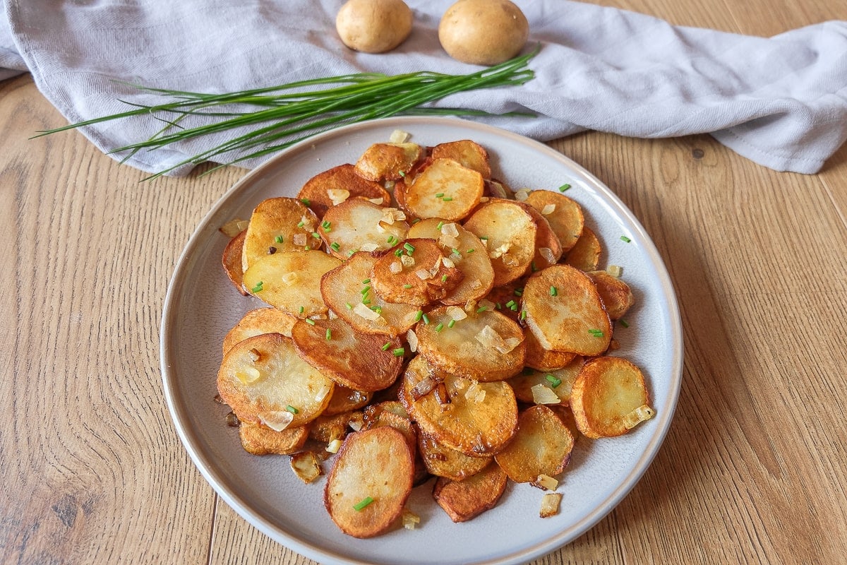 plate of german fried potatoes on wooden table with chives and potatoes behind
