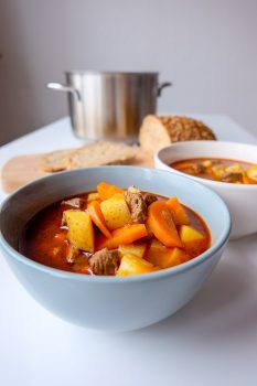 blue bowl of hungarian goulash soup on white table with bread and silver pot behind