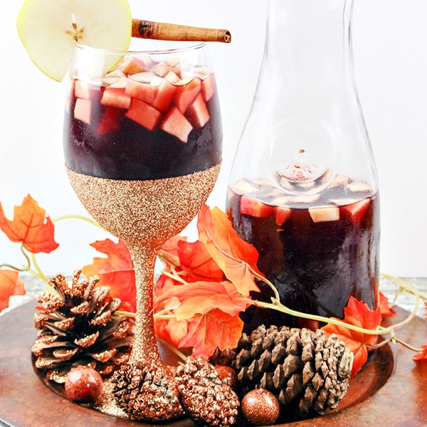 Pitcher of dark red sangria with apple cubes and wine glass with gold decor and sangria in front.