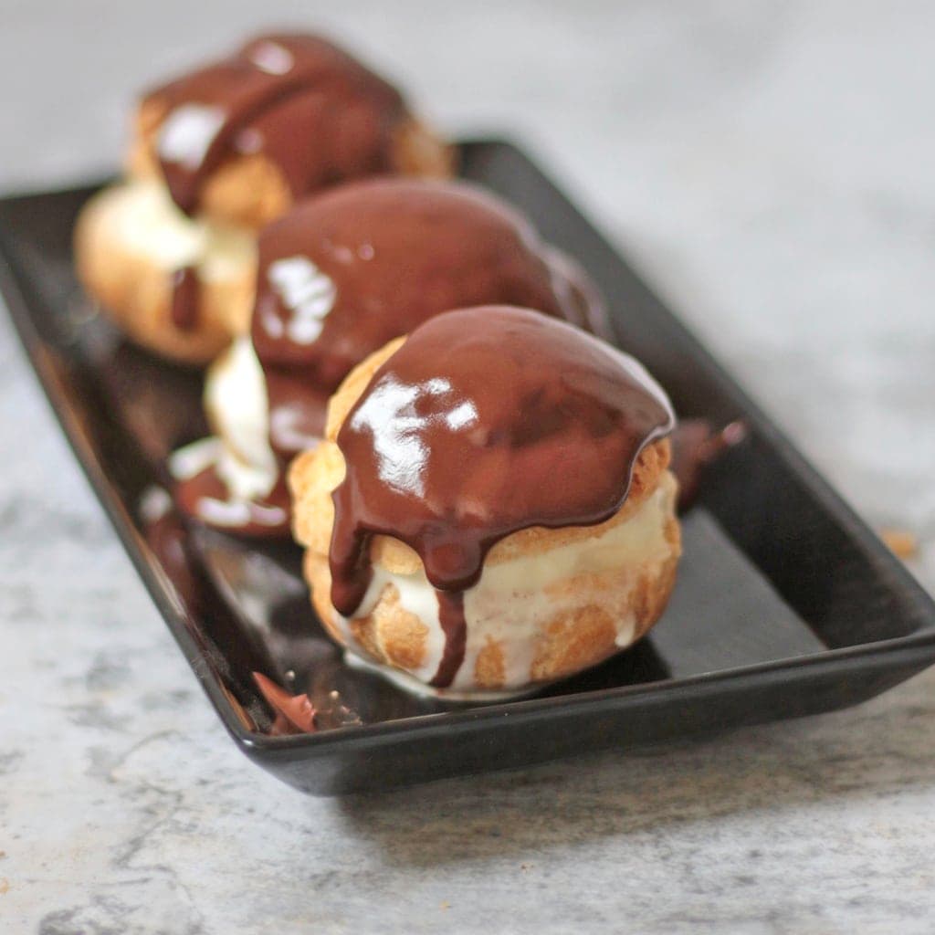 Cream puffs with chocolate drizzle on black plate.