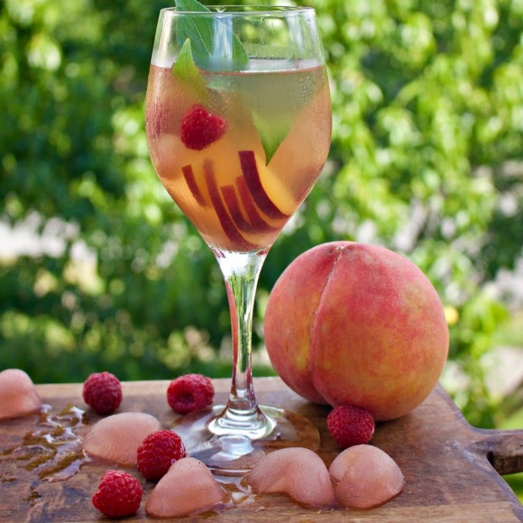 sangria with peach slices and raspberry in wine glass with whole peach next to it.