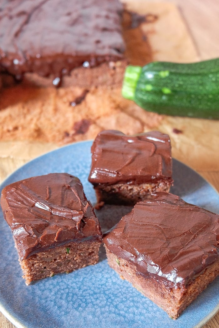 squares of cake with chocolate icing on blue plate with green zucchini behind