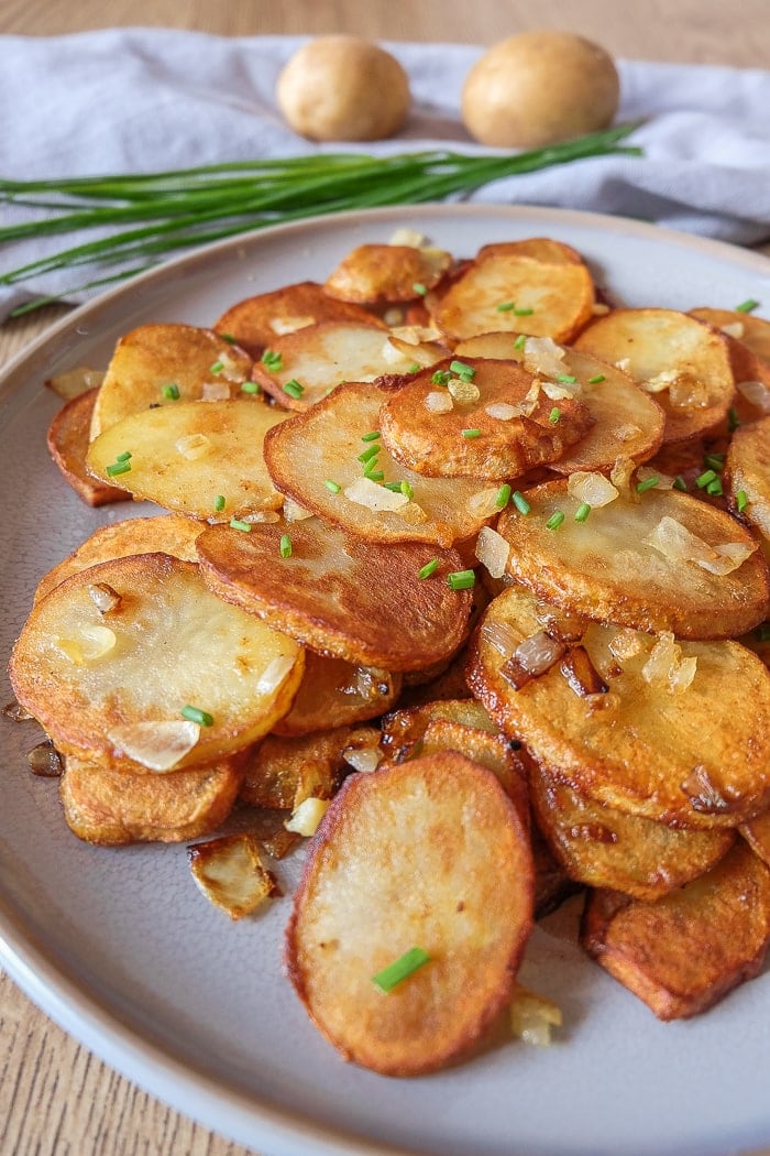 close up shot of fried potatoes on grey plate with potatoes and chives behind