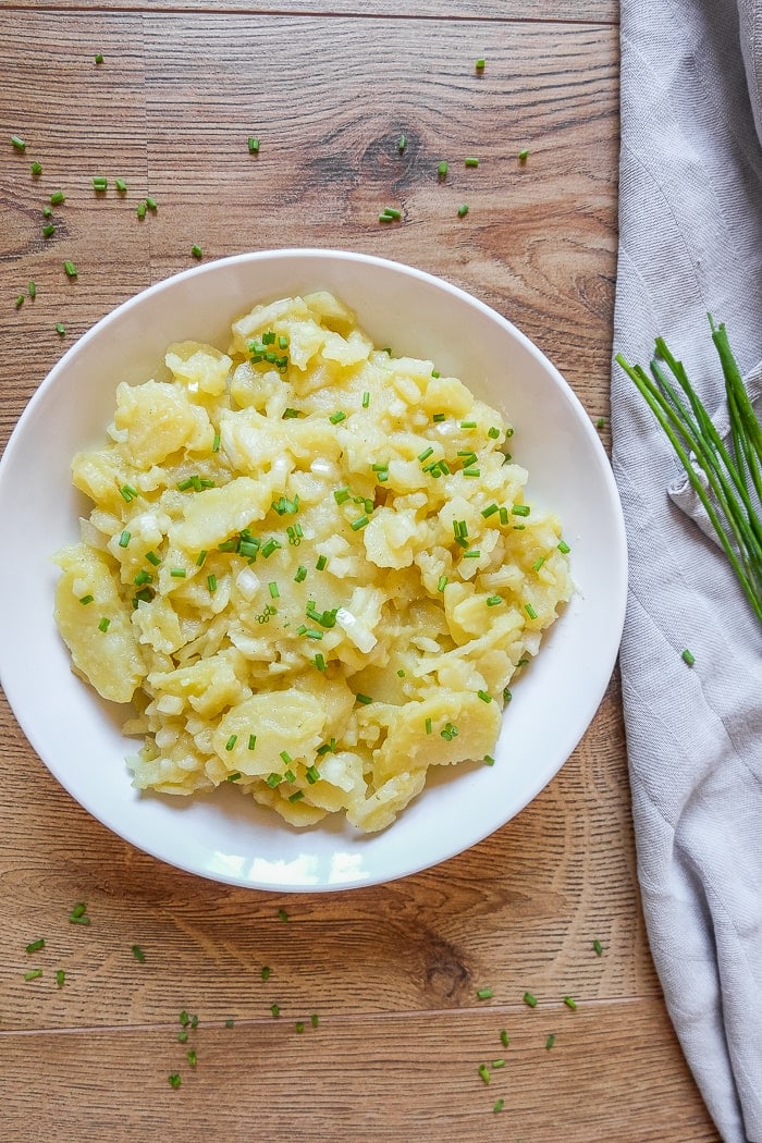 white bowl of potato salad beside grey cloth with green chives beside