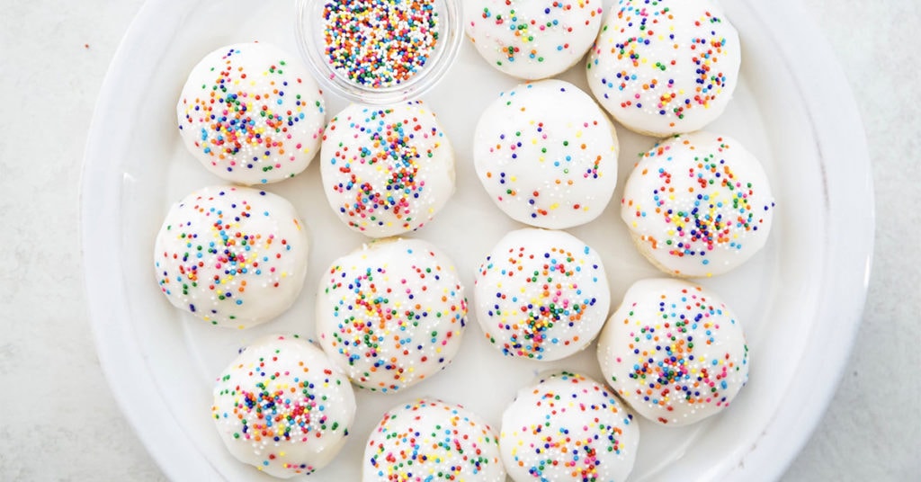 round anise cookies with white glaze and sprinkles on top on white plate.