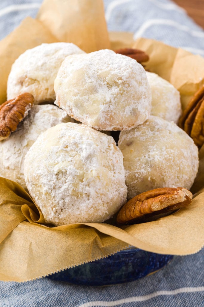 small round cookies dusted in icing sugar with pecans next to it in tin with parchment paper. 