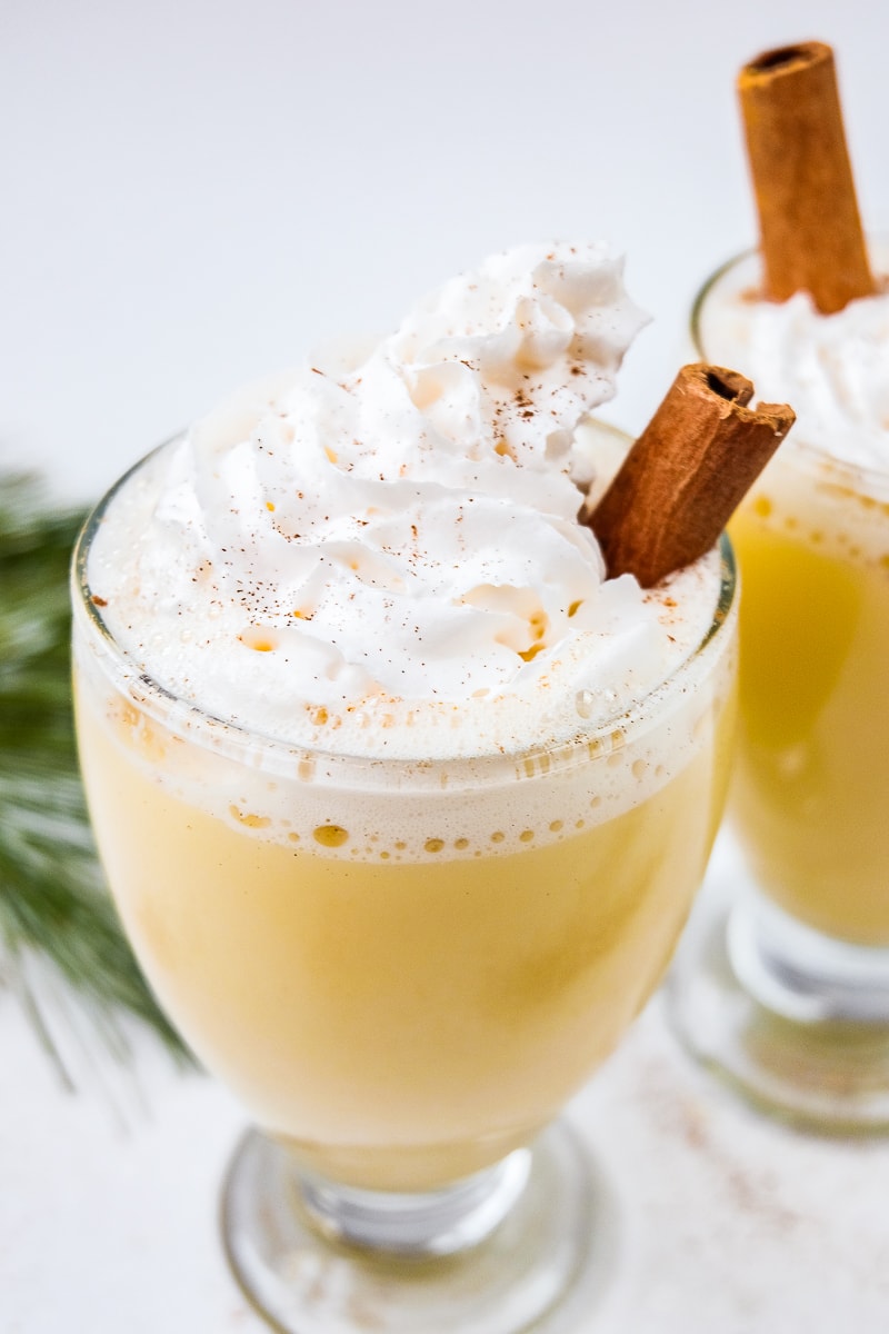 two glasses of egg punch topped with whipping cream with cinnamon stick sticking out