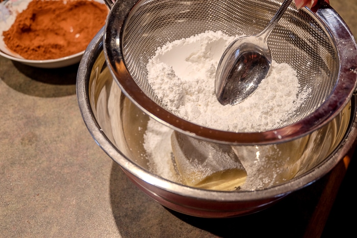 spoon sifting icing sugar into metal mixing bowl of marzipan
