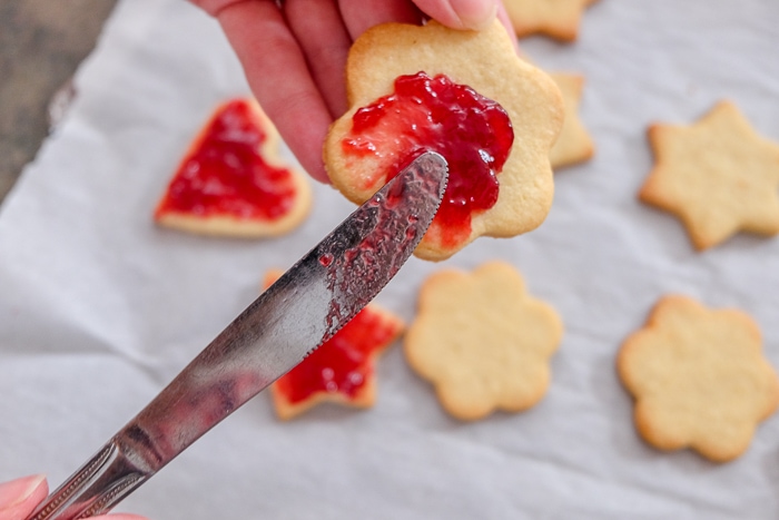 butter knife spreading red jam on linzer cookie