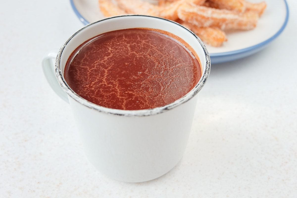 white mug of hot chocolate with plate of churros behind on white counter top