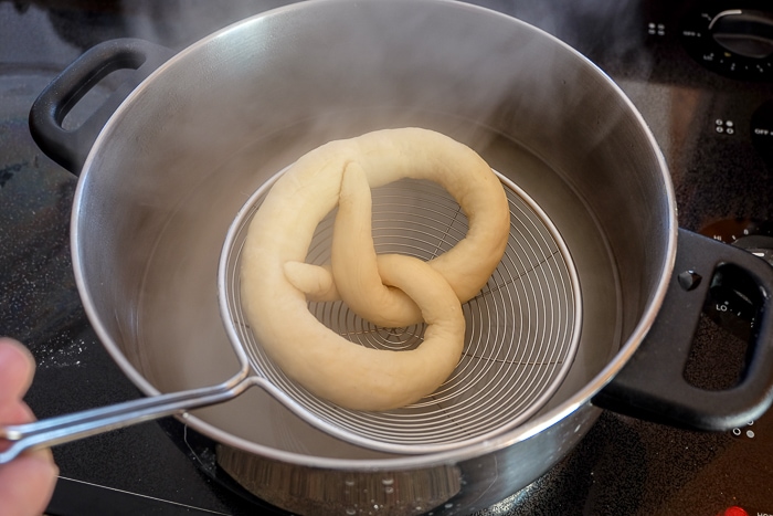 raw german pretzel dunking into pot of boiling water with metal strainer spoon