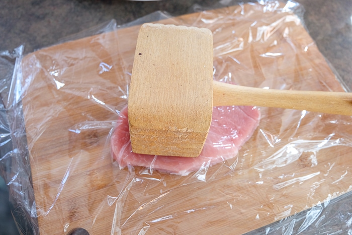 wooden meat hammer sitting on pork cutlet under plastic wrap on wooden cutting board