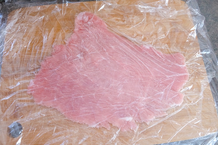 thinly pounded piece of pork under cling wrap on wooden board