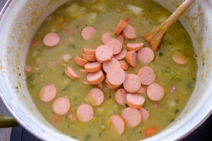 chopped weiner sausage in split pea soup in pot on stove with wooden spoon