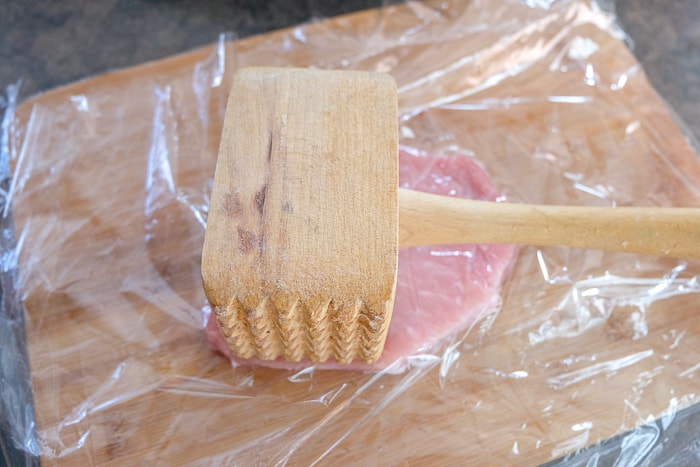 wooden meat hammer laying on pork chop under plastic wrap