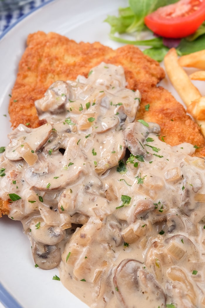 breaded schnitzel covered in mushroom sauce on plate with chopped parsley and fries