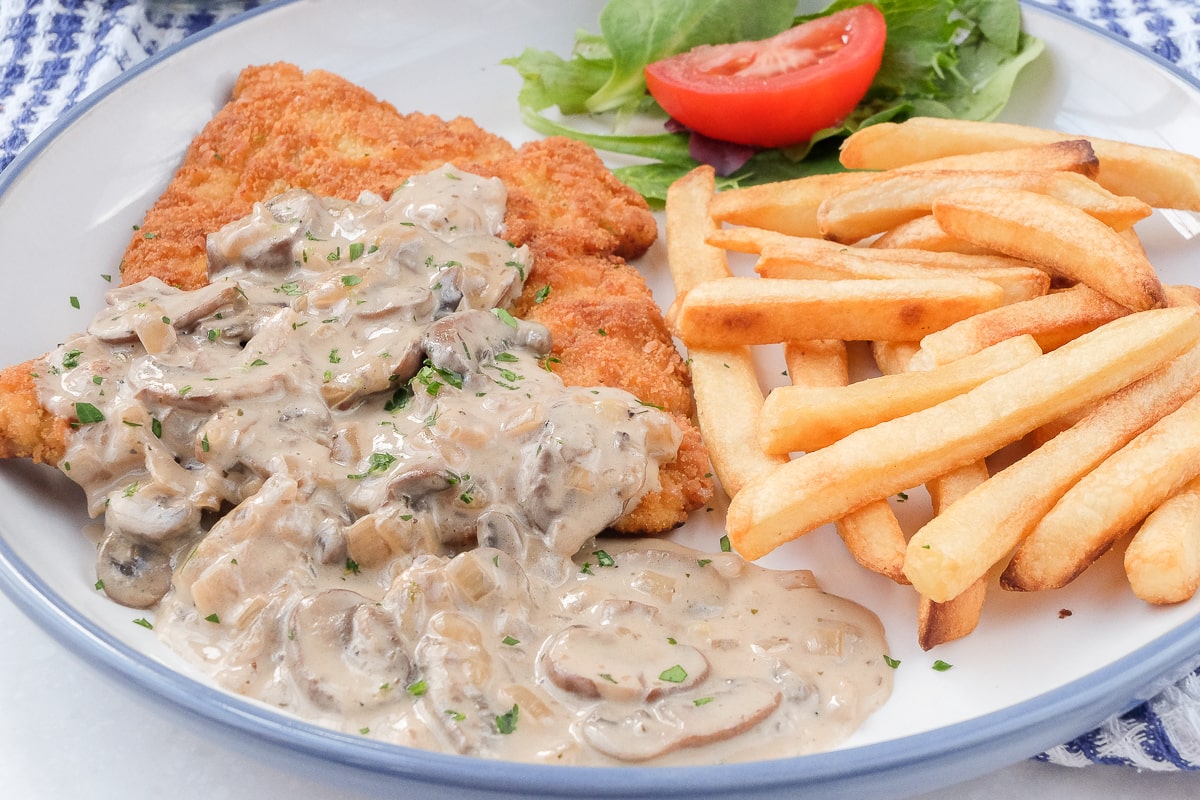 breaded schnitzel covered in mushroom sauce on plate with fries and small salad behind
