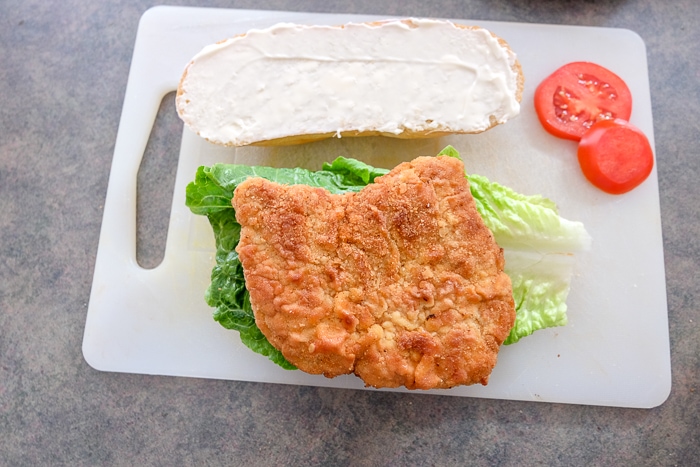 breaded schnitzel on bun with lettuce and tomatoes on cutting board