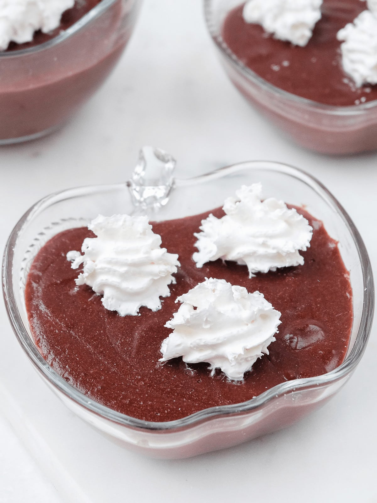 three glass bowls with chocolate pudding and whipping cream on top on white marble background