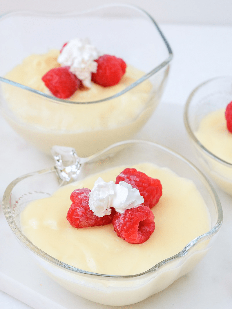 three clear bowls with yellow vanilla pudding in it and raspberries and whipping cream on top