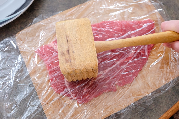 wooden hammer pounding beef between cling wrap on wooden board