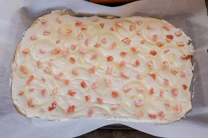raw flammkuchen with cream onion and bacon on top on baking pan
