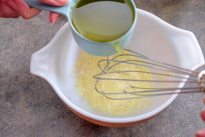 olive oil being added into white mixing bowl with silver whisk inside stirring