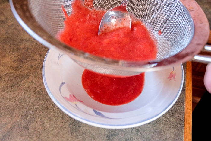 strawberry puree pushed with silver spoon through metal sifter into white bowl below