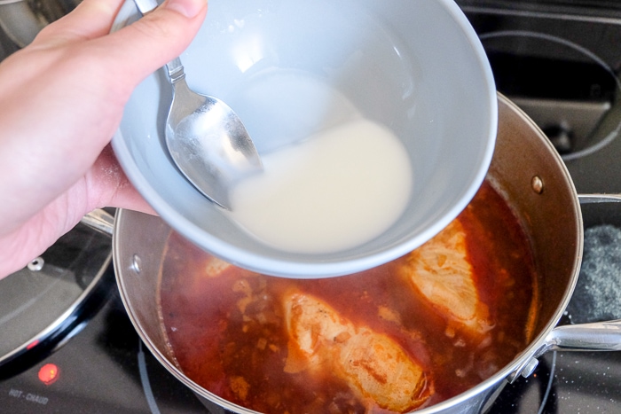 pouring corn starch into red chicken broth on stove top