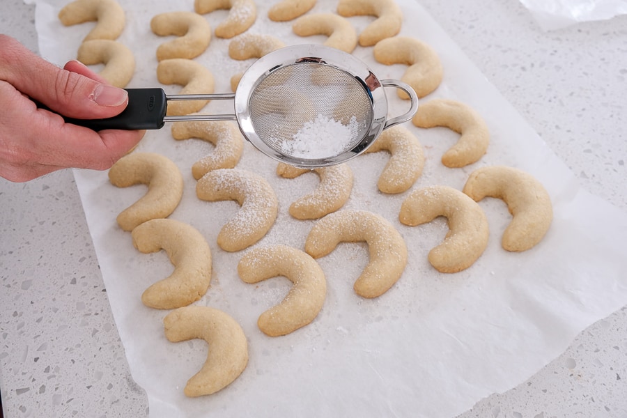 silver strainer sprinkling powdered sugar on crescent cookies in counter