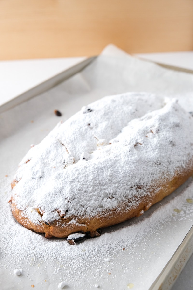 german stollen loaf covered in icing sugar sitting on parchment paper pan with wood behind.