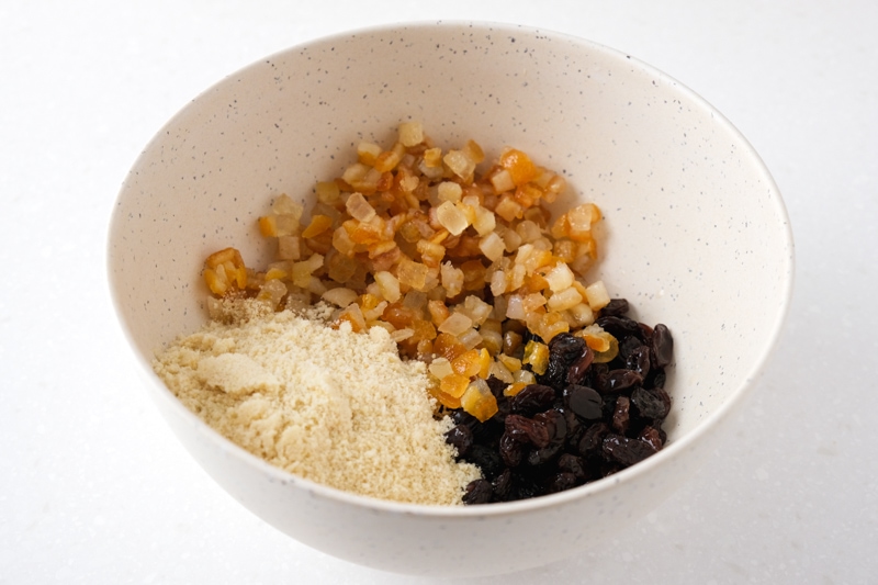 beige mixing bowl filled with raisins candied peels and breadcrumbs on white counter.