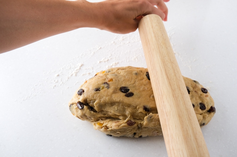 wooden rolling pin in hand pressing down on ball of stollen dough on countertop.