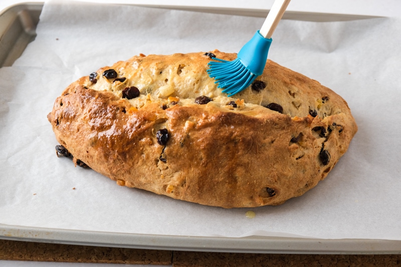 blue silicone tipped brush putting melted butter on baked loaf of german stollen sitting on parchment paper.