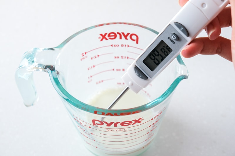 hand holding digital thermometer sticking out of glass pyrex cup sitting on counter. 