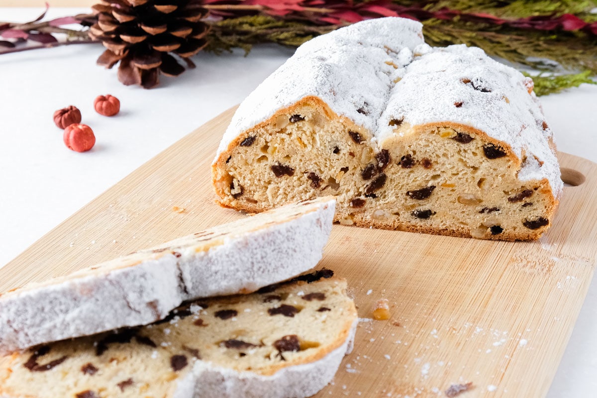 a half loaf of stollen on wooden board with red berries beside and two slices in front.