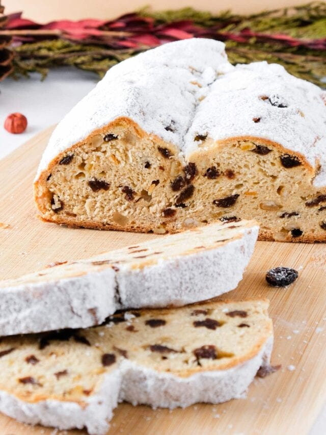 loaf of german stollen with two slices cut on wooden board with christmas branches behind.