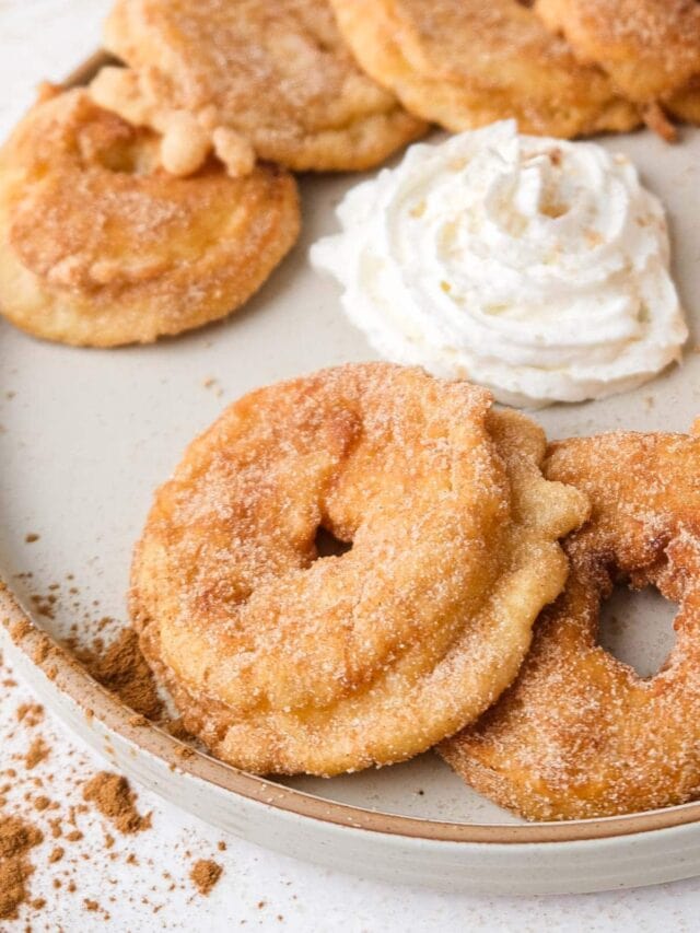 fried apple ring with cinnamon and sugar on plate with other rings and whipped cream behind.