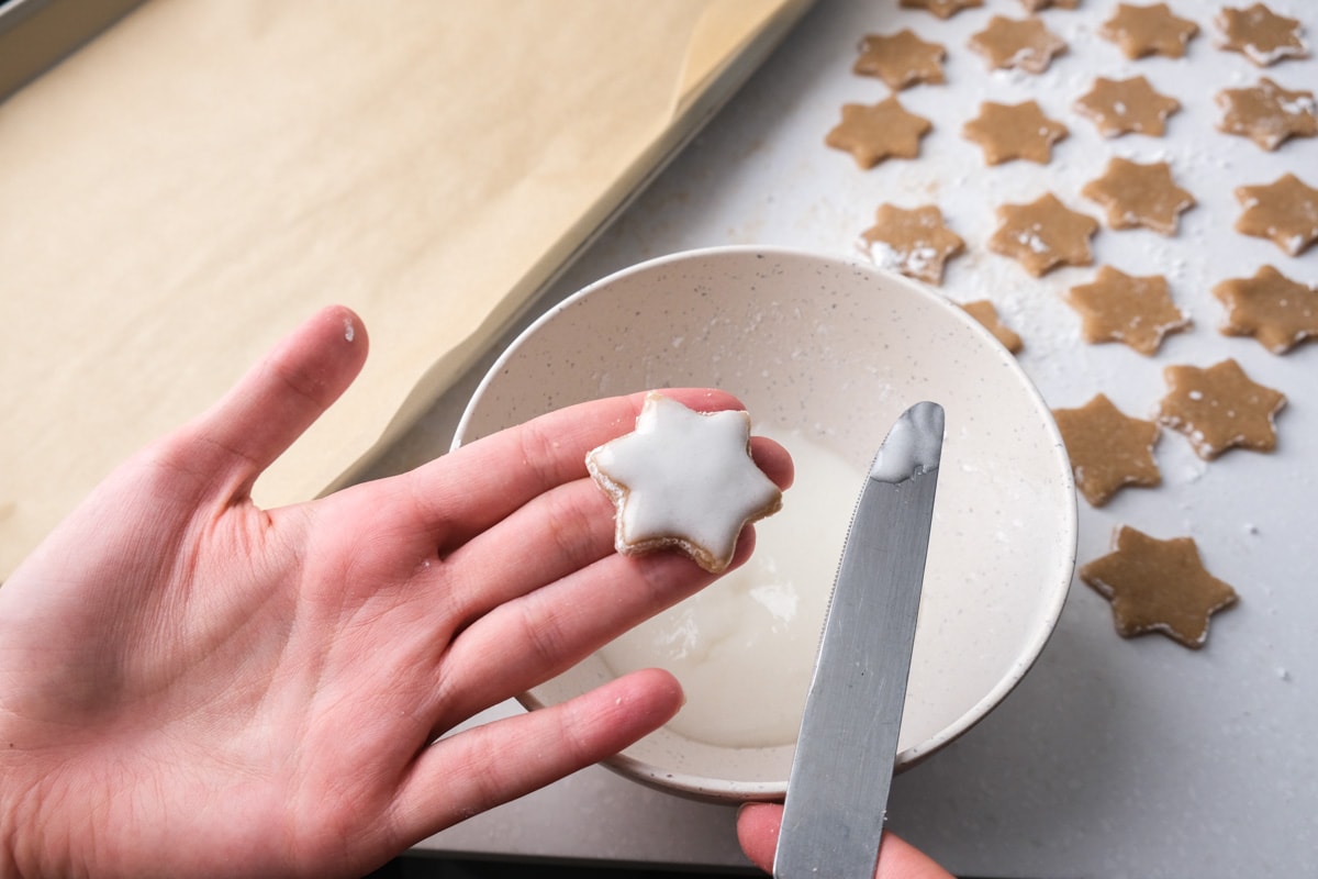 hand holding cinnamon star cookie with white icing on top with more cookies behind.