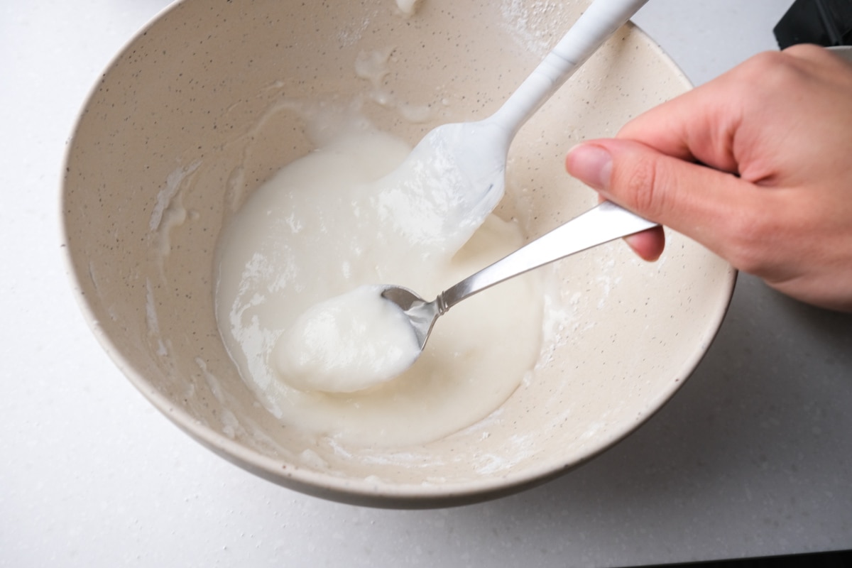 silver spoon testing icing in white mixing bowl on counter.