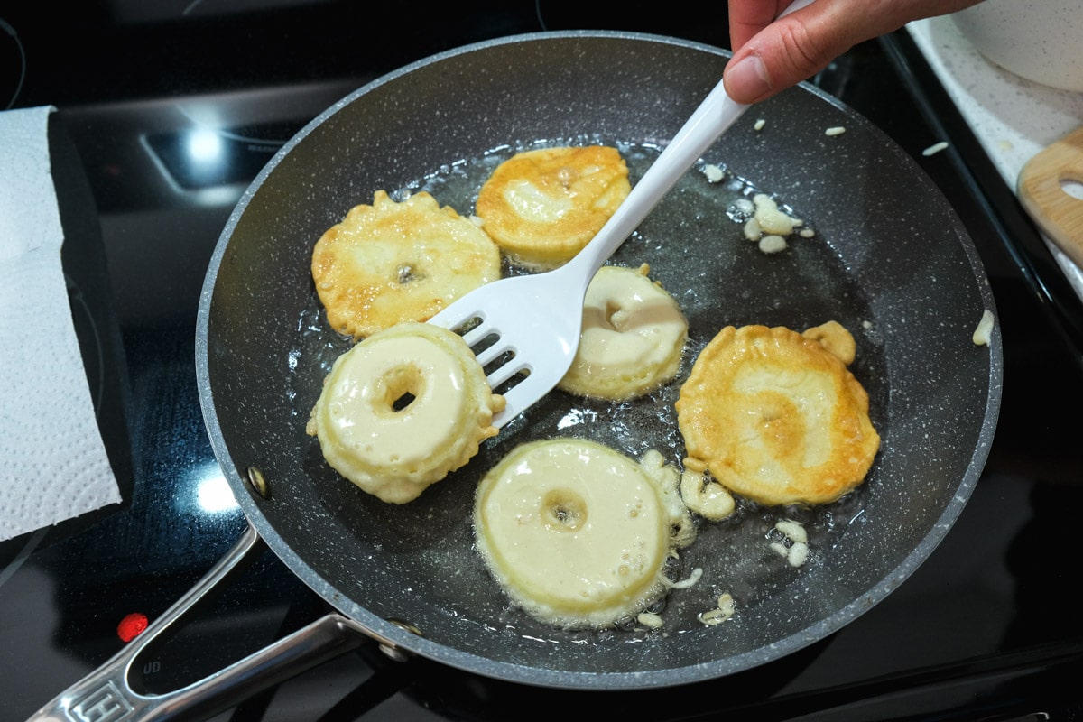 white spatula flipping fried apple rings in frying pan full of oil.