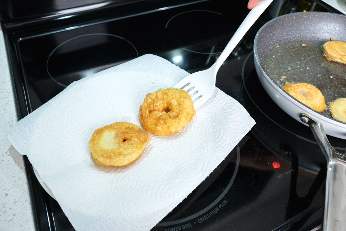 white flipper moving fried apple rings onto paper towel plate on stove top.