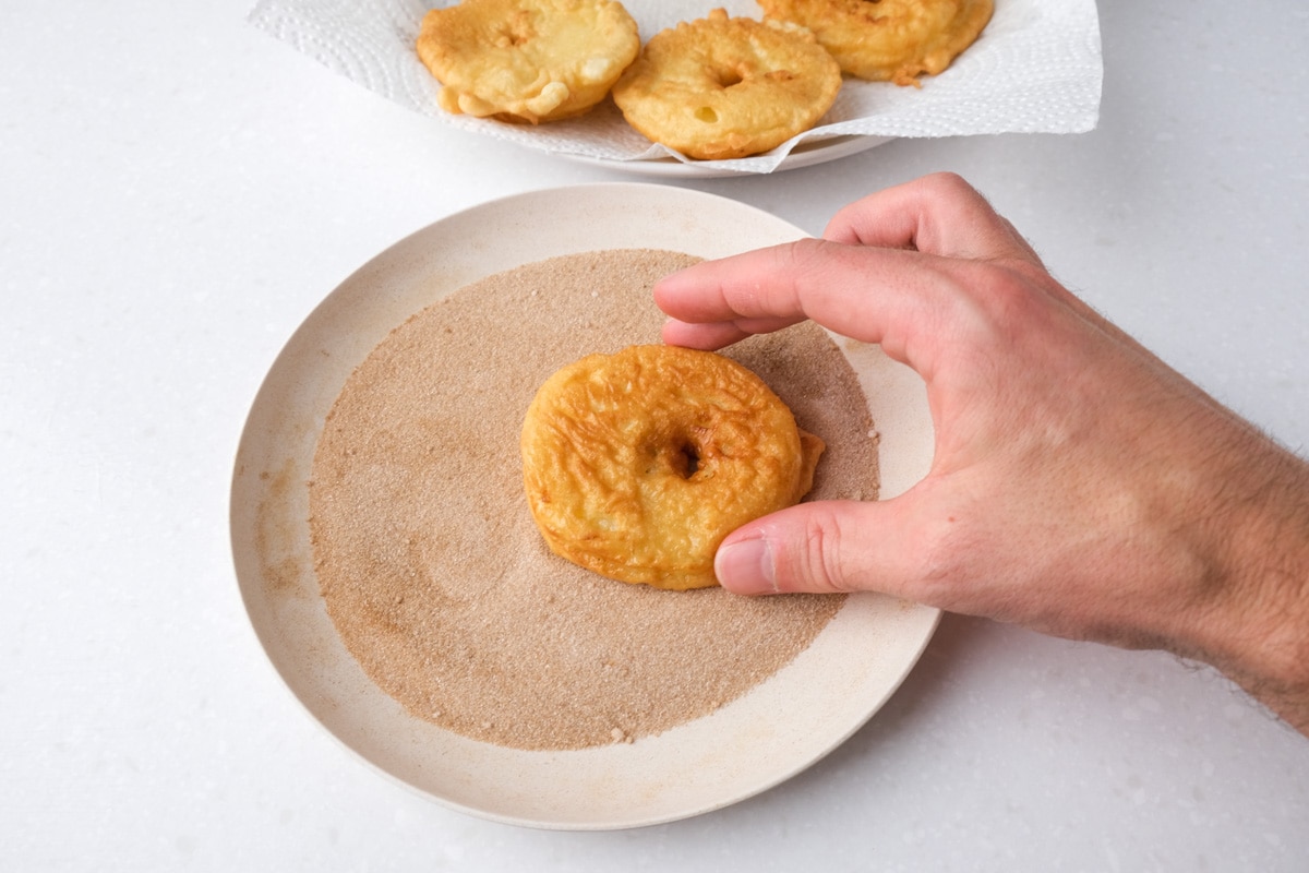 hand holding fried battered apple ring over plate of cinnamon and sugar on white counter.
