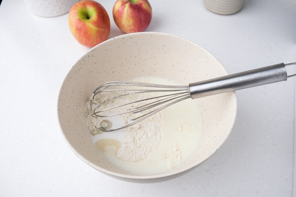 silver whisk in white bowl of batter with red apples behind.