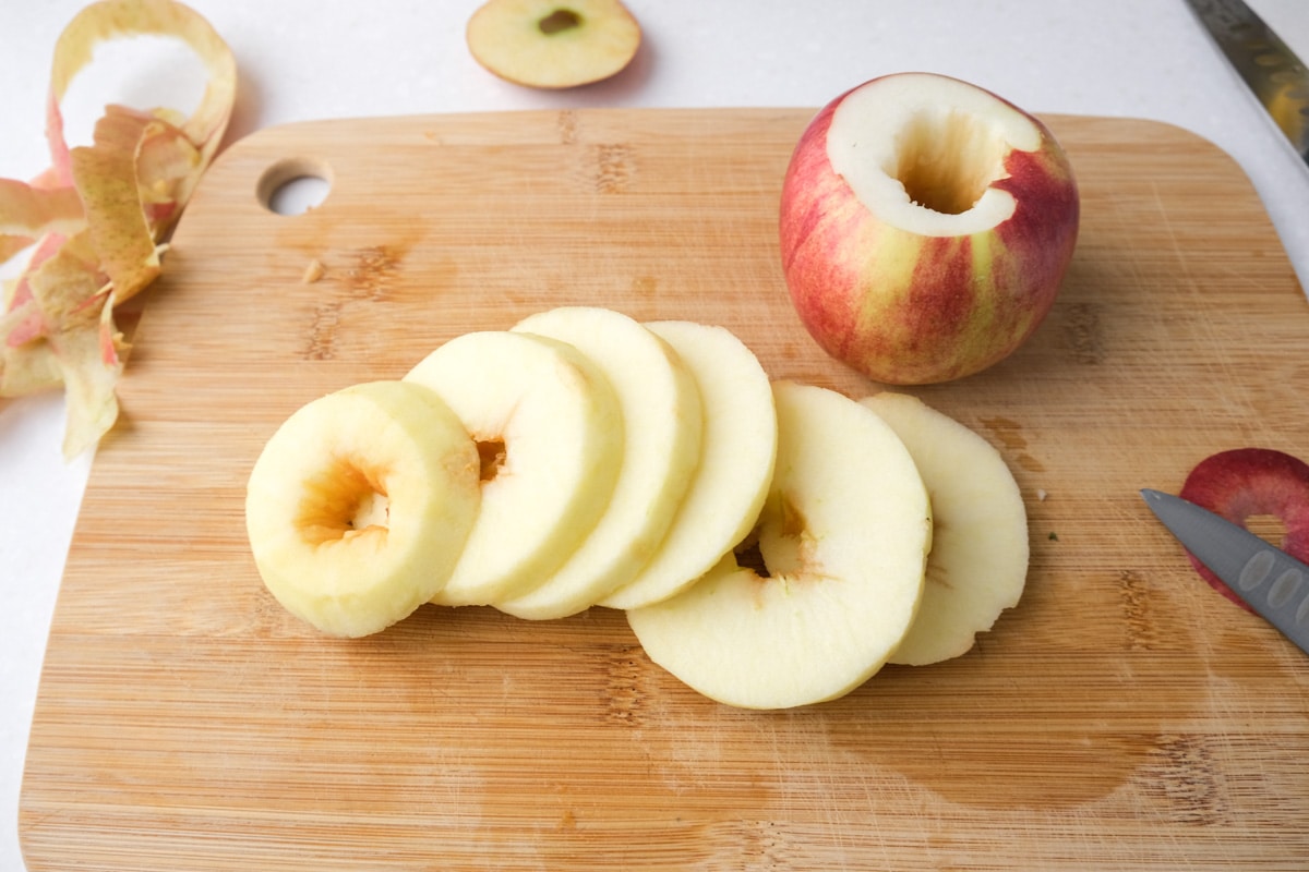 peeled apple cut into slices with whole apple behind all sitting on wooden cutting board.