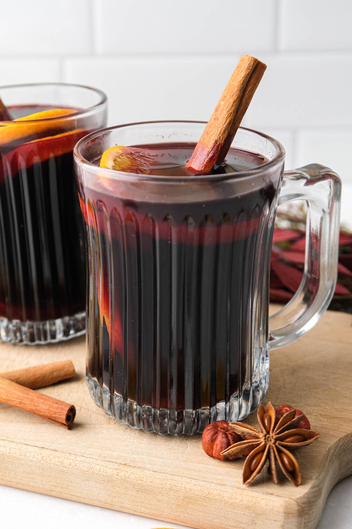 clear mug of mulled red wine with cinnamon stick sticking out sitting on wooden board with spices arounc.