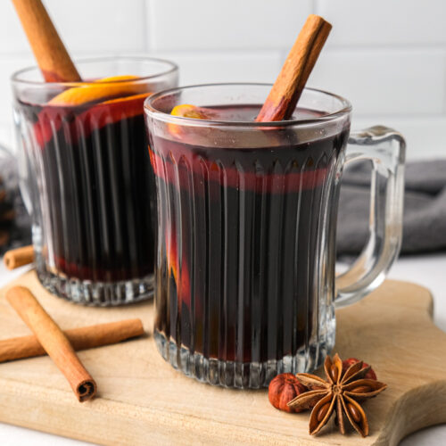 two clear mugs of mulled wine with cinnamon sticks sitting on wooden board.