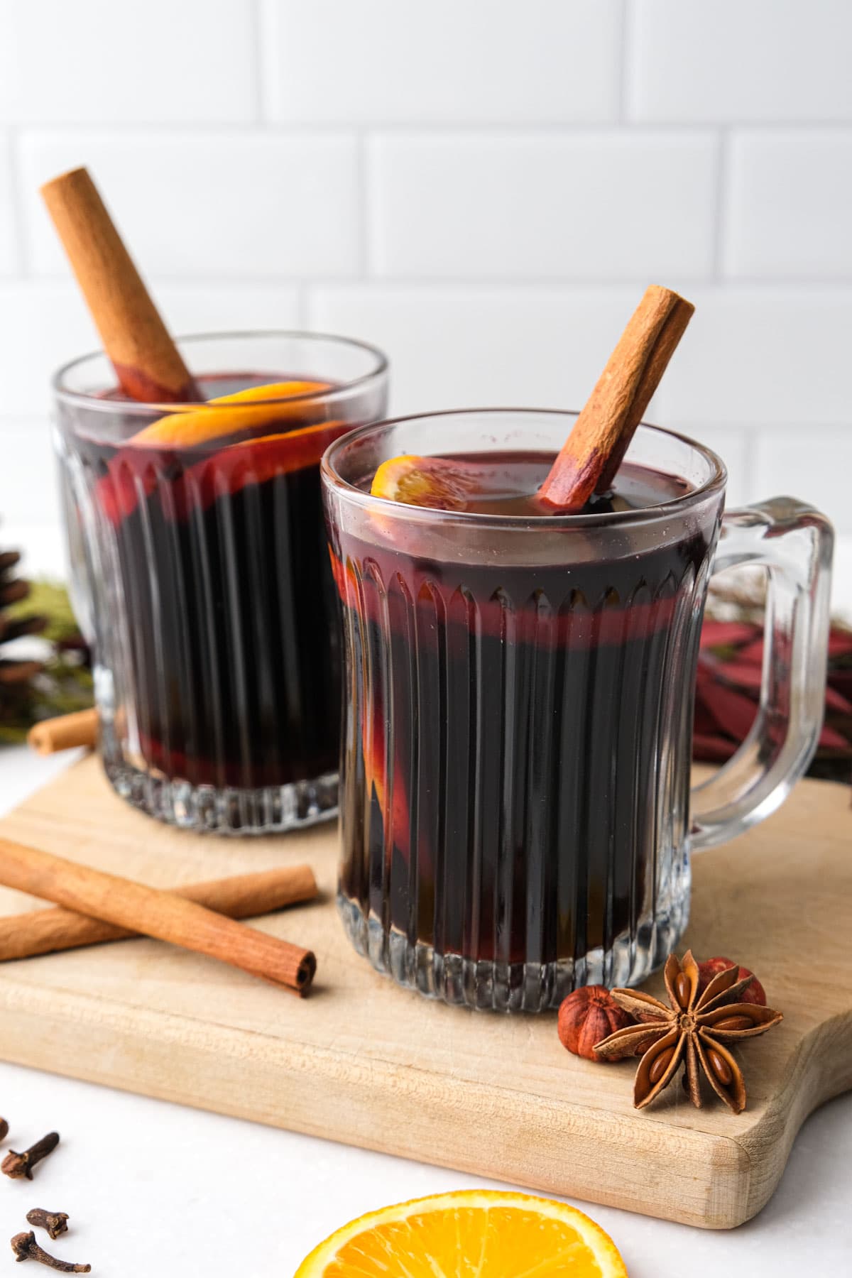 two mugs of mulled red wine on wooden board with spices around on white counter.