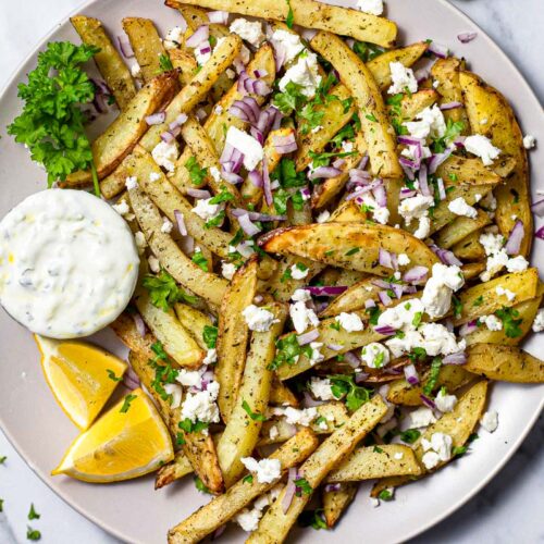 plate of greek fries covered in feta and onion on counter top with parsley beside.